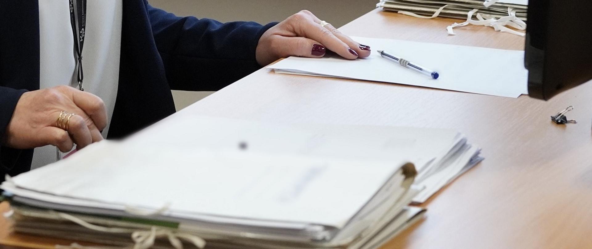 Na biurku leżą teczki z dokumentami. Przy biurku siedzi osoba. 