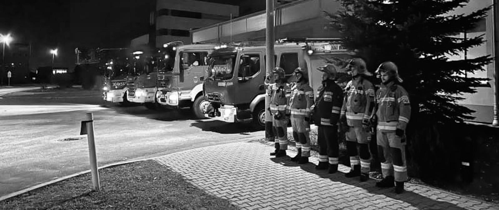 Strażacy stoją przed jednostką w szeregu. W tle wozy bojowe.