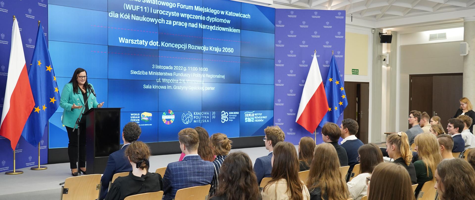 W sali konferencyjnej publiczność siedzi na krzesłach. Na scenie na tle ścianki w mównicy wiceminister Małgorzata Jarosińska-Jedynak. Obok flagi PL i UE.