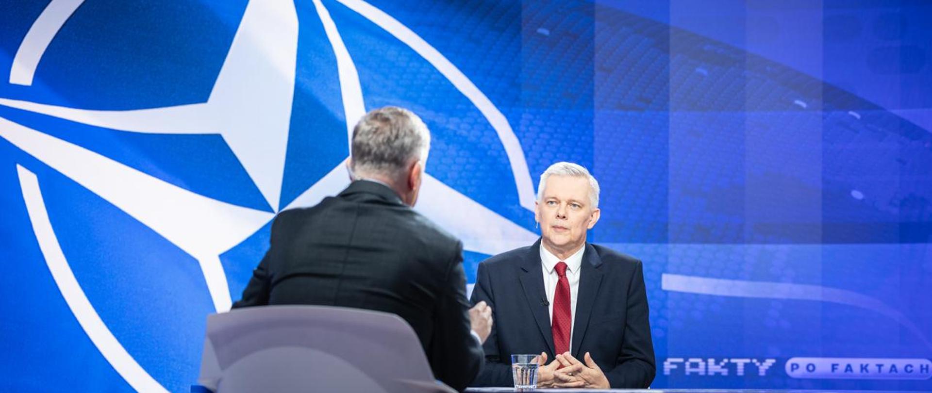 Na zdjęciu przy stole siedzą: minister Tomasz Siemoniak (po prawej, przodem do obiektywu) oraz redaktor Grzegorz Kajdanowicz (po lewej, tyłem do obiektywu). Na stole widać szklankę z wodą. W tle niebieska flaga NATO z białym logo Sojuszu. W prawym dolnym rogu zdjęcia napis "FAKTY PO FAKTACH".