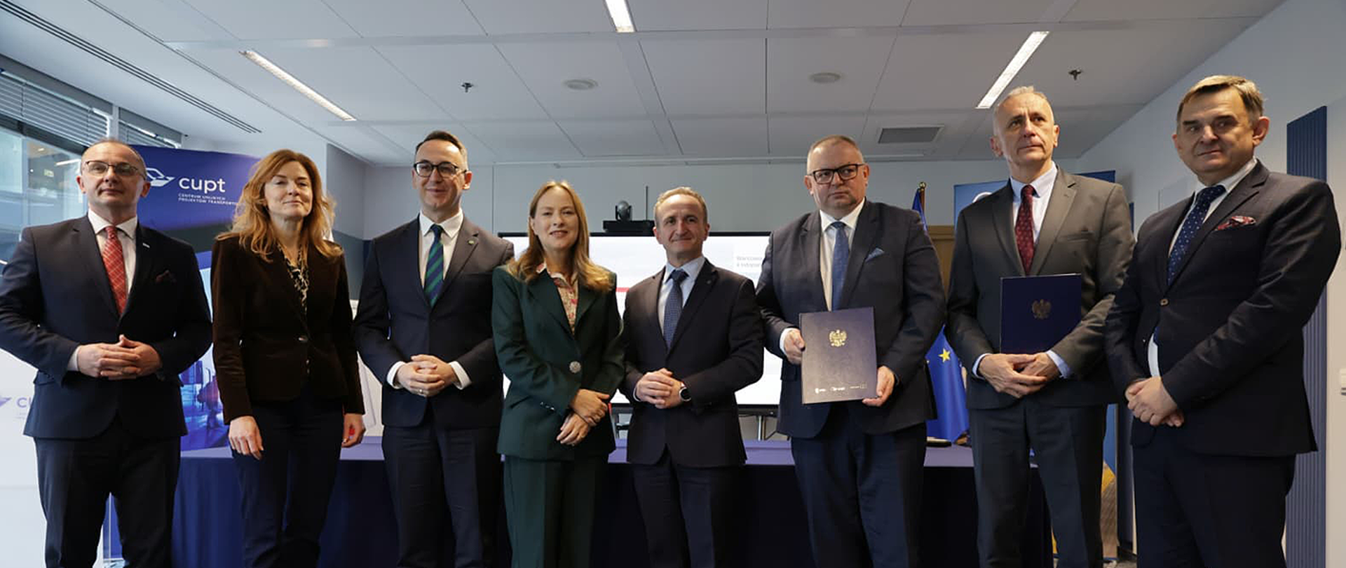Grupa ośmiu osób pozuje do wspólnego zdjęcia po podpisaniu umów o dofinansowanie zakupu nowych pociągów dla Śląska. W centrum stoi minister funduszy i polityki regionalnej Katarzyna Pełczyńska-Nałęcz oraz przedstawiciele województwa śląskiego, Kolei Śląskich i CUPT. W tle widoczny jest baner Centrum Unijnych Projektów Transportowych oraz flaga Unii Europejskiej