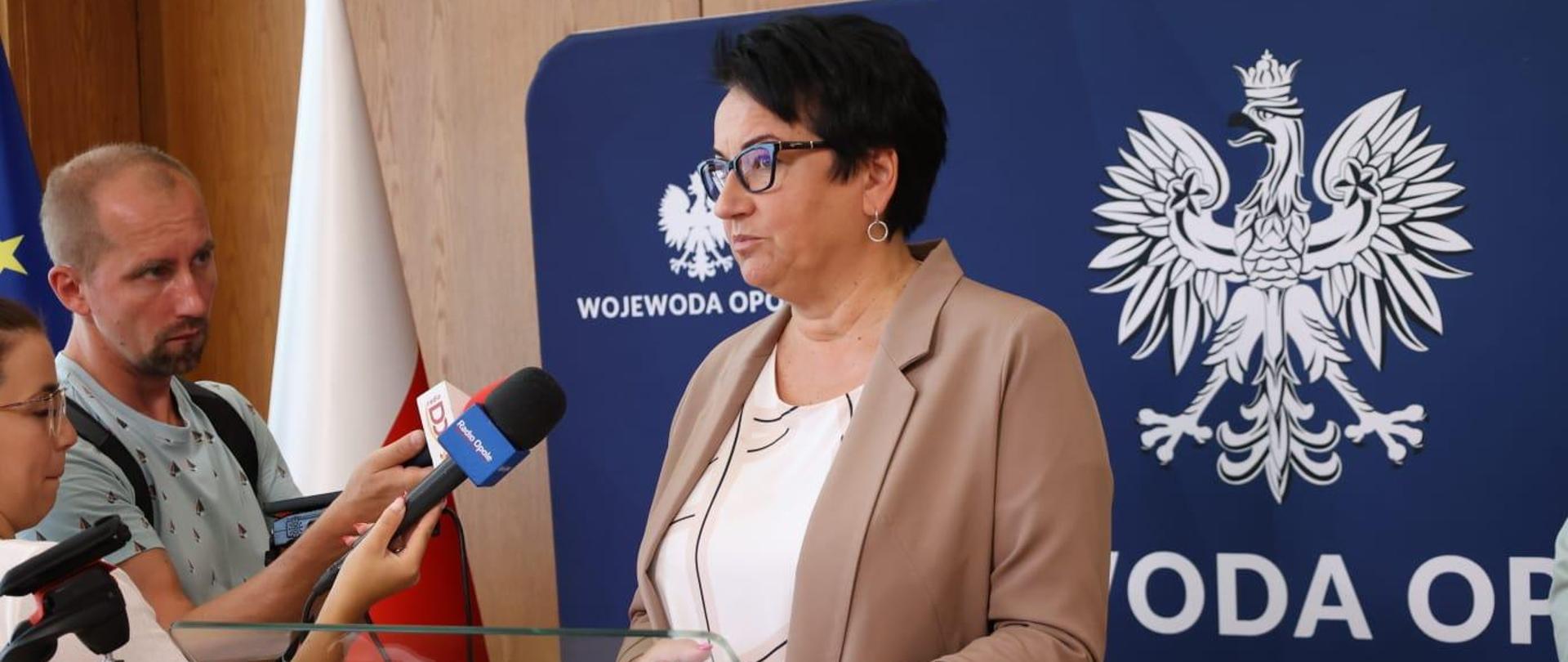 Wojewoda Opolski Monika Jurek stoi przed mównicą. Zdjęcie zrobione jest od pasa w górę. Po lewej stronie stoją dziennikarze z mikrofonami, do których mówi Wojewoda. 