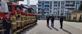 Komendant główny Państwowej Straży Pożarnej w obecności kierownictwa Komendy Wojewódzkiej Państwowej Straży Pożarnej stoi przed strażakami stojącymi w rzędzie.
