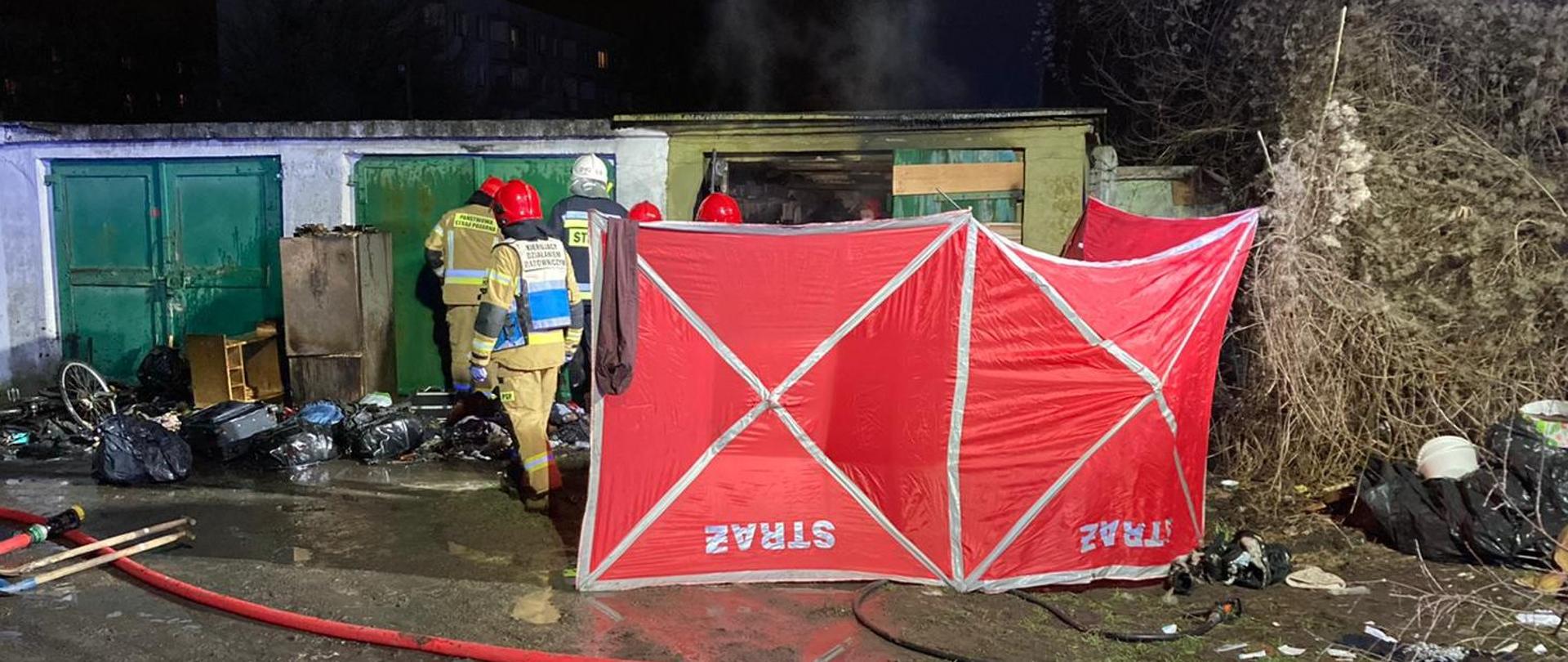 Zdjęcie wykonane w porze nocnej przy sztucznym oświetleniu. Na pierwszym planie znajduje się parawan strażacki osłaniający pożar garażu w ciągu garaży konstrukcji murowanej.
Za parawanem widoczni strażacy PSP i OSP. Przed parawanem linia gaśnicza oraz linia szybkiego natarcia. W tle widoczne budynki wielorodzinne.