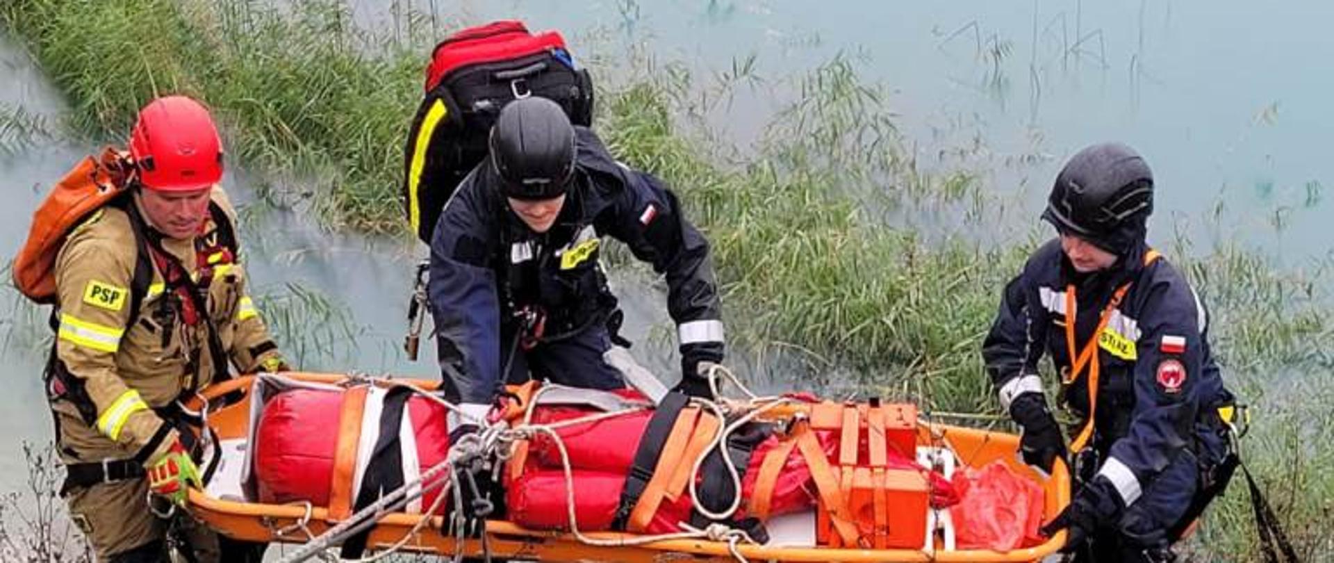 Zdjęcie wykonane podczas ćwiczeń na terenie zalanej przez wodę kopalni piachu w miejscowości Wierzbica. Na zdjęciu widać trzech strażaków ze specjalistycznej grupy ratownictwa wysokościowego. Strażacy przy pomocy lin i noszy wchodzą wraz z osobą poszkodowaną znajdującą się na noszach pod strome zbocze. Osobę poszkodowaną pozoruje manekin wykonany z tworzywa sztucznego w kolorze czerwonym. Strażacy ubrani są w specjalne kombinezony do ratownictwa wysokościowego w kolorze niebieskim i piaskowym. Na głowach posiadają kaski. Dodatkowo jeden z nich, znajdujący się przy noszach na plecach ma założoną torbę medyczną do udzielania pierwszej pomocy.