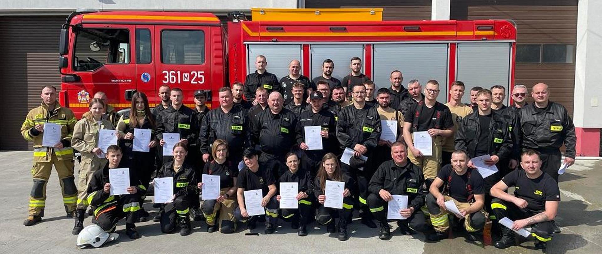 na tle samochodu pożarniczego grupa druhów OSP po zdanym egzaminie pozująca do zdjęcia. pożarniczego