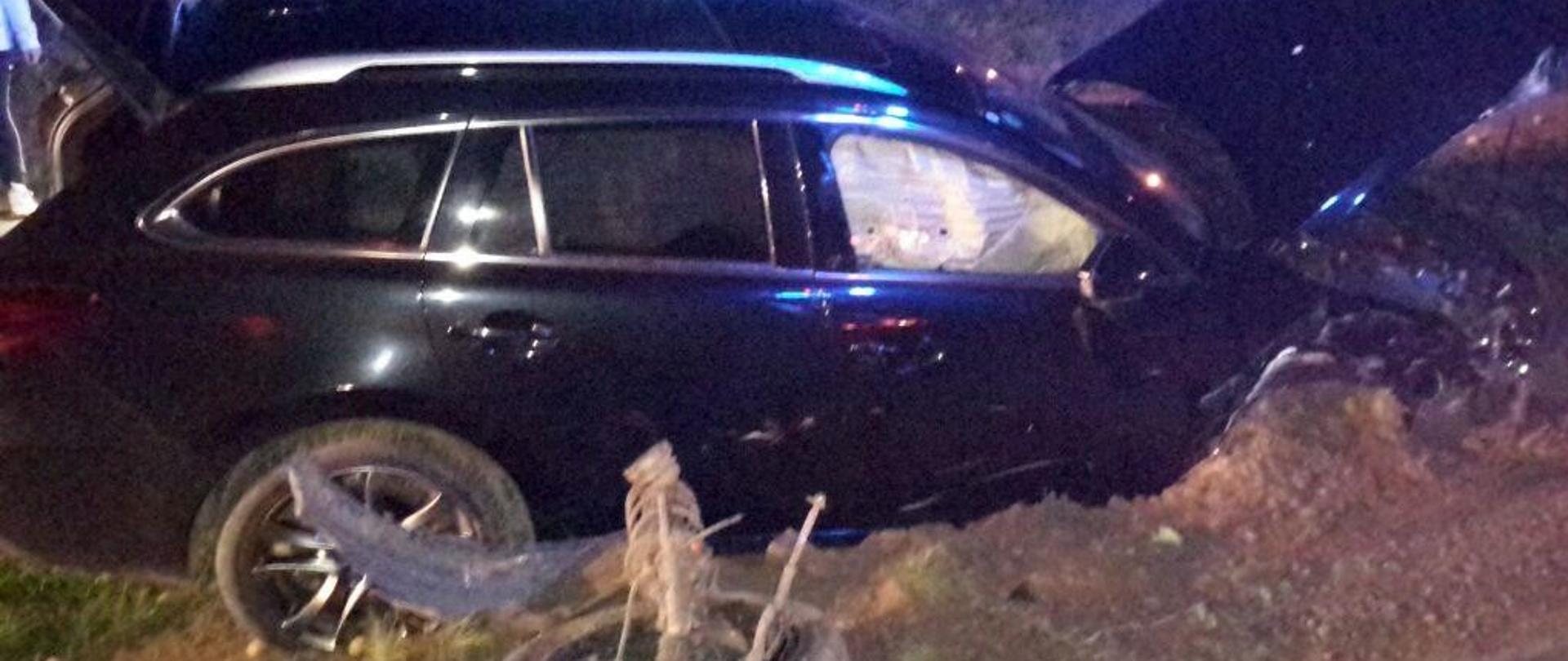 Uszkodzony czarny samochód, częściowo zakopany w ziemi. Przed nim oderwane koło. Za samochodem strażacy uczestniczący w działaniach. W tle fragment ogrodzenia posesji.