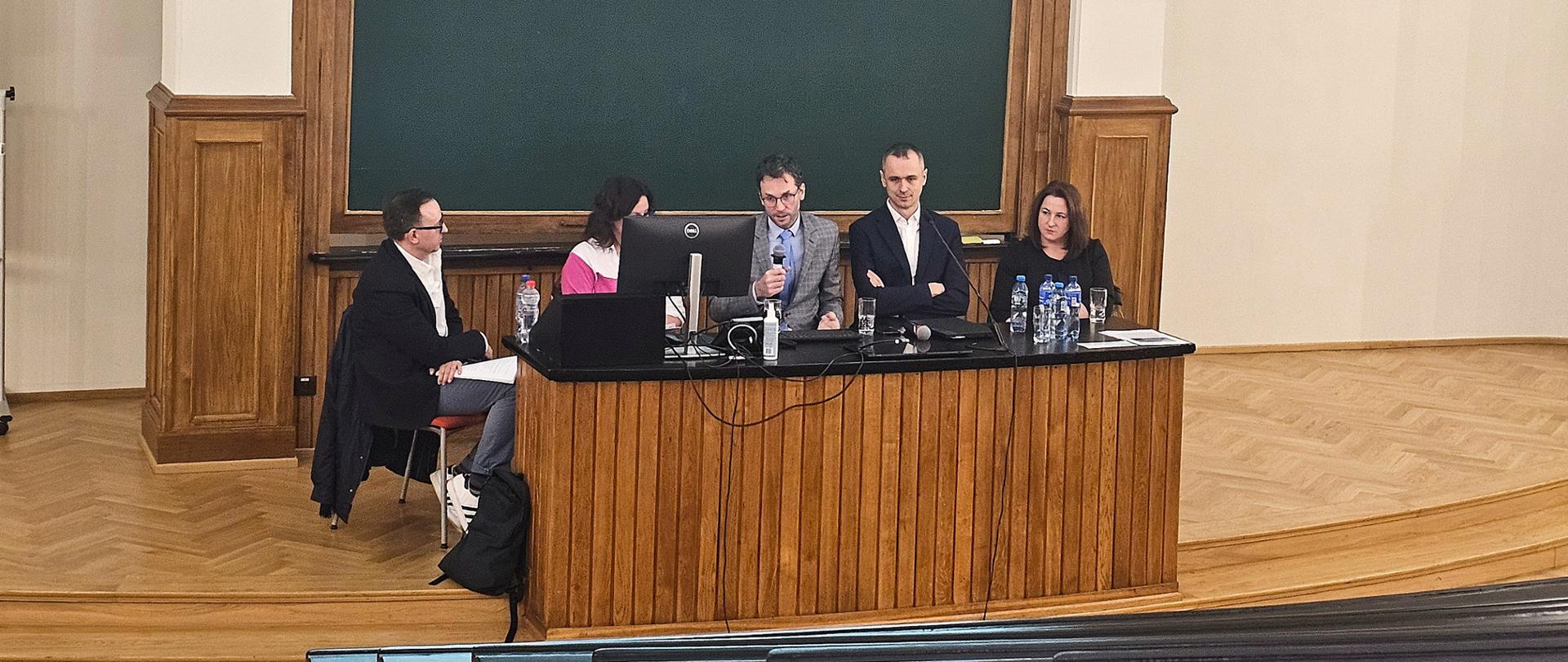 Pięć osób siedzi za stołem prezydialnym na sali wykładowej, jedna z nich mówi do mikrofonu,