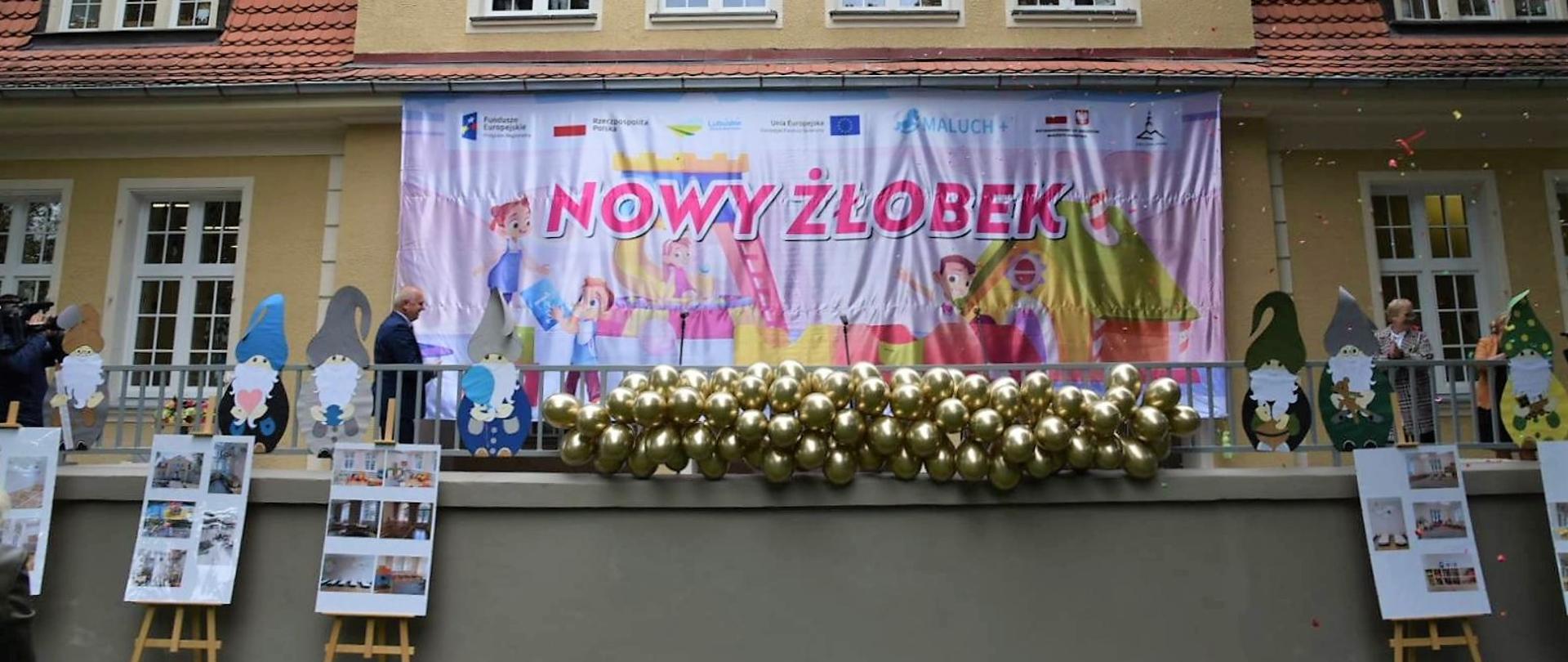 Na budynku, w wejściu powieszony duży i kolorowy baner z napisem "Nowy Żłobek". Przy nim stoi wojewoda i inni goście 
