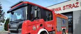 Samochód pożarniczy Scania ustawiony przed garażem komendy, nad nim napis Państwowa Straż Pożarna.
