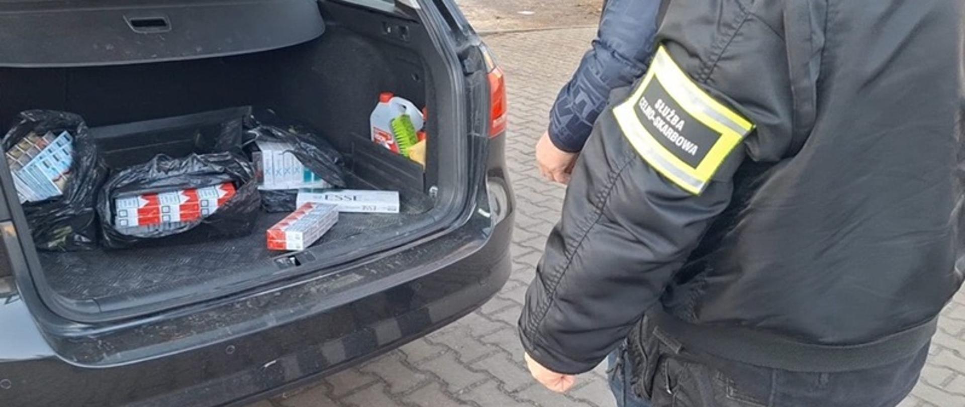 Osoby stojące przed samochodem z otwartym bagażnikiem, w którym leżą paczki papierosów. 