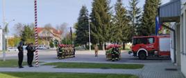 Na placu przed Jednostka Ratowniczo Gaśnicza nr 1 stoją strażacy w dwóch szpalerach naprzeciw siebie na uroczystej zbiórce. Pomiędzy szpalerami stoi dowódca. Na maszt uroczyście wciągana jest biało czerwona flaga. Strażacy ubrani są w stroje bojowe. Za drugim szpalerem widoczny jest samochód gaśniczy.