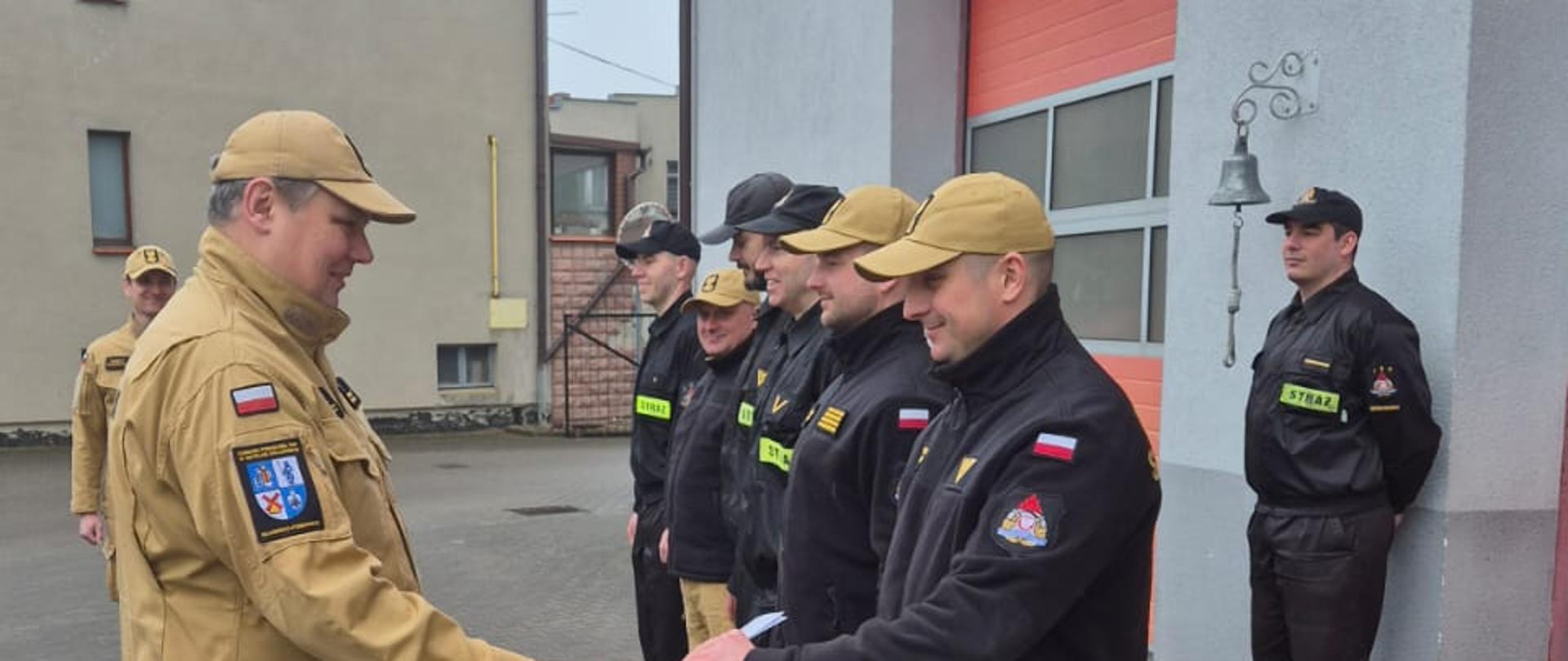 Funkcjonariusze w ubraniach koszarowych na placu komendy przed garażami, Komendant powiatowy brygadier Marcin Mazurkiewicz w ubraniu służbowym wręcza list gratulacyjny funkcjonariuszowi Przemysławowi Szmelter.