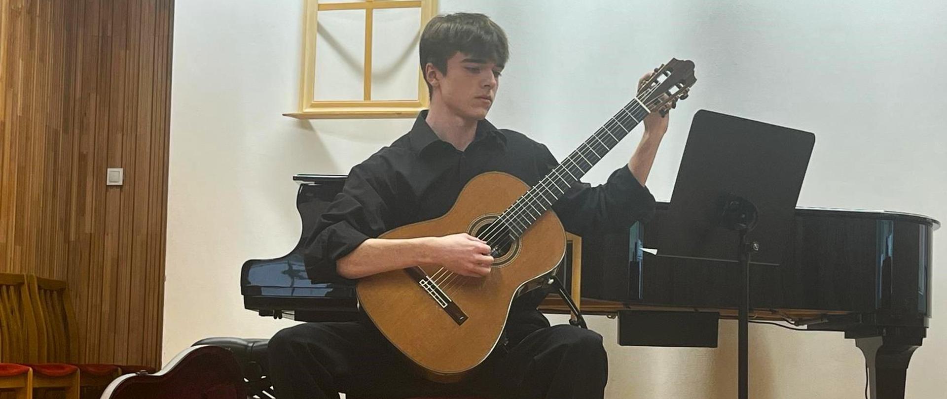 Na zdjęciu Karol Kubiec podczas występu, siedzi na krześle, na scenie, gra na gitarze. Na podłodze stoi otwarty futerał, z tyłu fortepian.