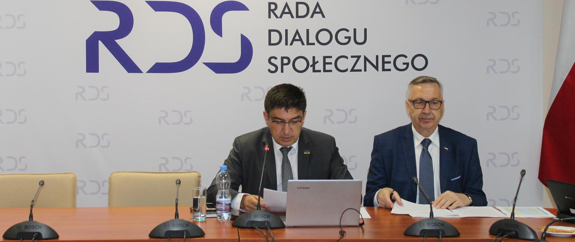 Na zdjęciu widnieje stół prezydialny za którym siedzą licząc od lewej strony" Przewodniczący Zespołu - Wojciech Ilnicki a obok wiceminister rodziny i polityki społecznej Stanisław Szwed. W tle osób siedzących widnieje baner z napisem Rada Dialogu Społecznego. Po prawej stronie zdjęcia stoi flaga Polski.