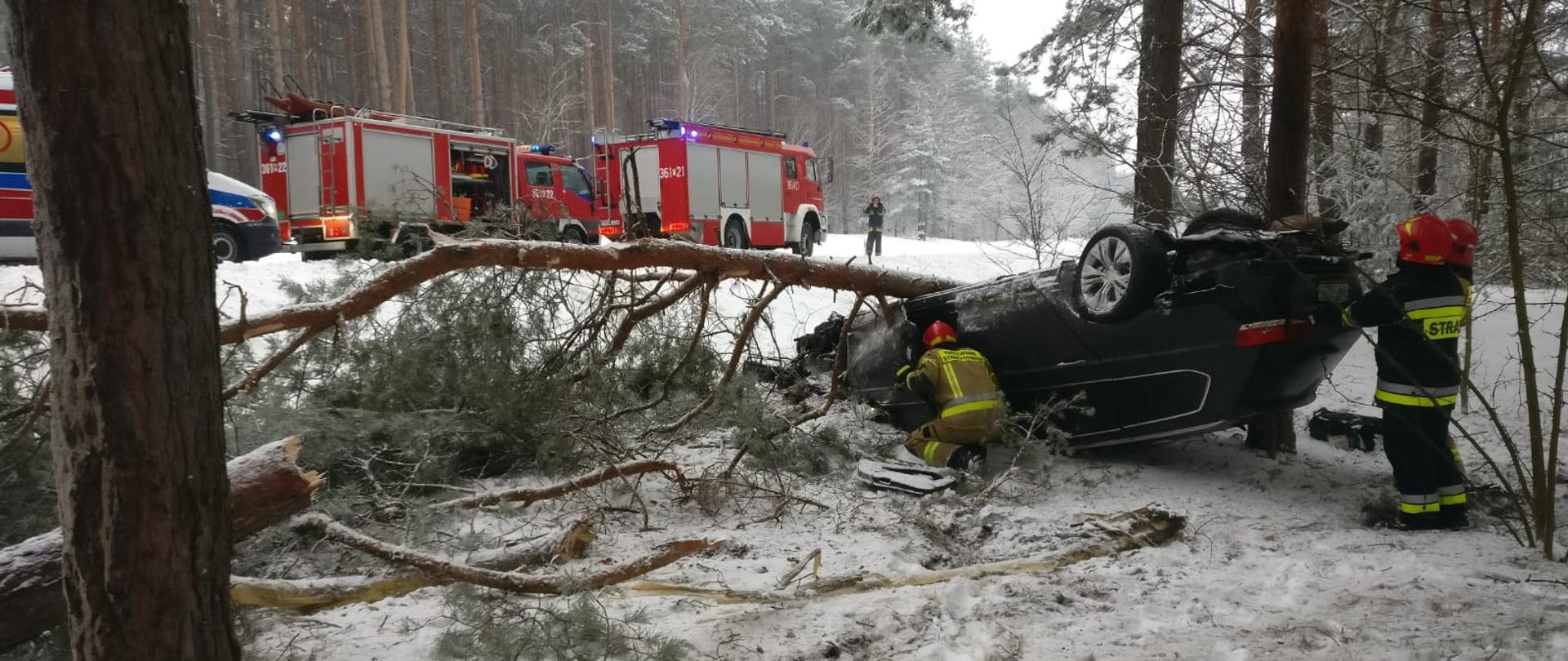 Zdjęcie przedstawia czarny samochód osobowy na dachu, znajdujący się poza pasem drogowym. Przy samochodzie stoi dwóch strażaków oraz trzeci pochylony. W oddali znajdują się dwa samochody pożarnicze oraz pojazd zespołu ratownictwa medycznego.