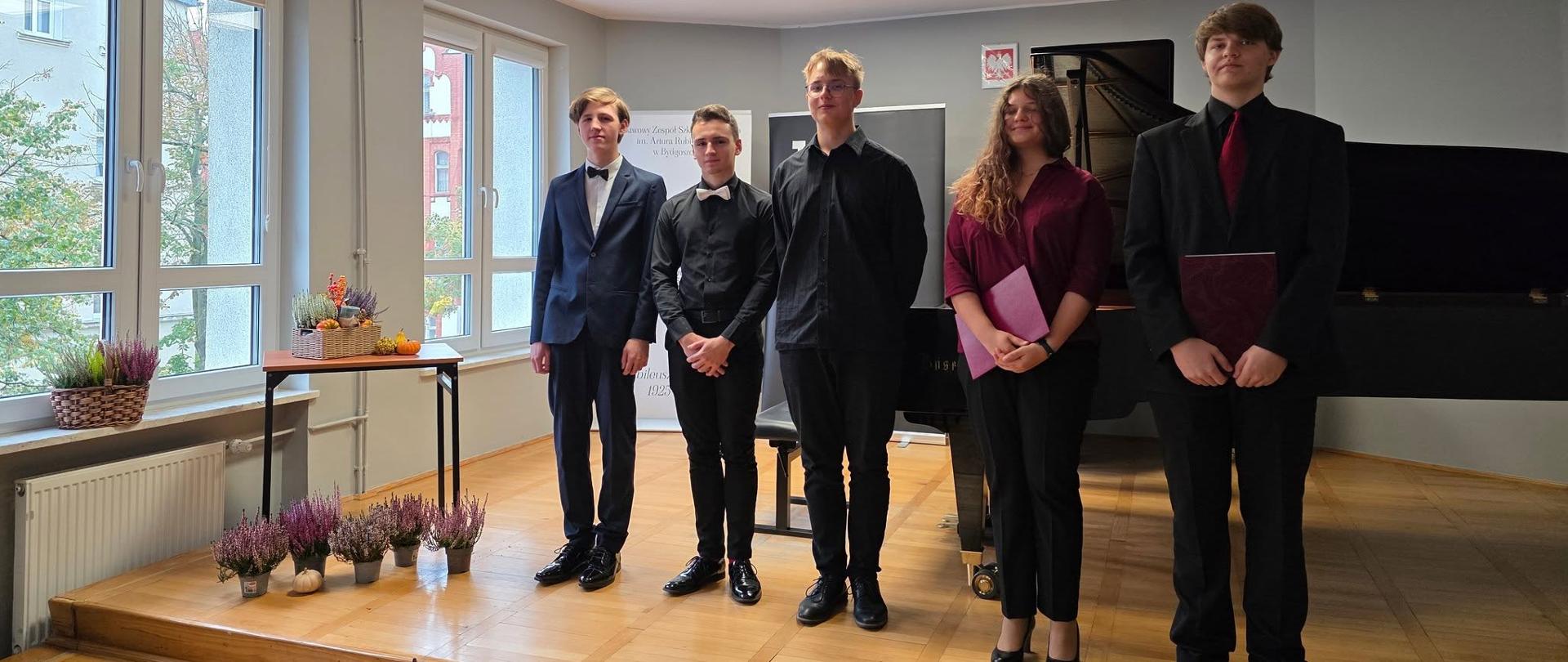 na zdjęciu na tle szarej ściany trzech mężczyn ubranych na czarno i jedna kobieta w długich brązowych włosach w czerwonej bluzce i czarnych spodniach obok stoją różowe kwiaty