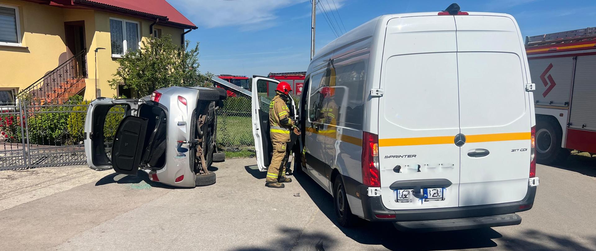Na zdjęciu dwa uszkodzone pojazdy. Samochód osobowy znajduje się na lewym boku przy ogrodzeniu posesji, natomiast bus znajduje się na poboczu drogi. Obok samochodu typu bus działania ratownicze prowadzi strażak w umundurowaniu specjalnym.
