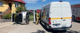 Na zdjęciu dwa uszkodzone pojazdy. Samochód osobowy znajduje się na lewym boku przy ogrodzeniu posesji, natomiast bus znajduje się na poboczu drogi. W środku samochodu typu bus działania ratownicze prowadzi strażak w umundurowaniu specjalnym.