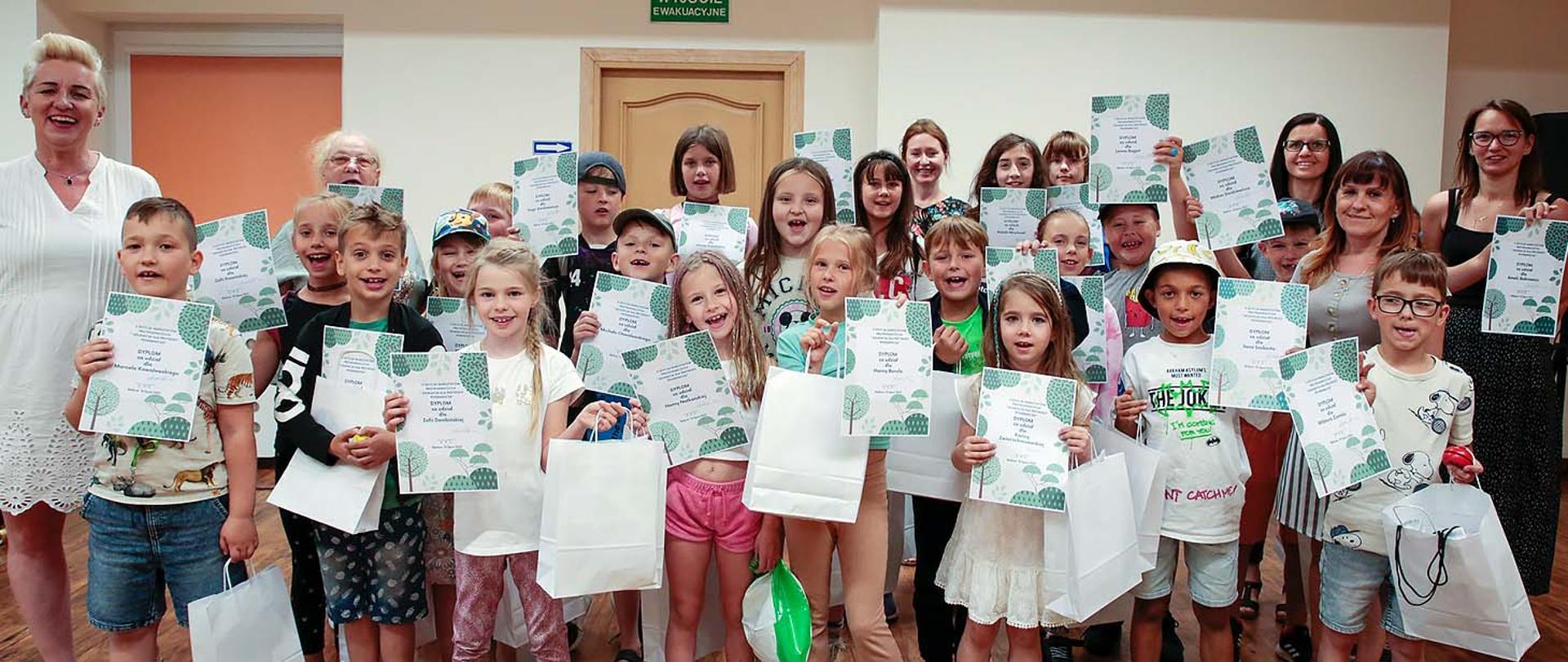 Na zdjęciu duża grupa dzieci i osób dorosłych stojąca w pomieszczeniu, dzieci trzymają w rękach dyplomy i papierowe torby.