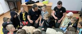 Grupa dzieci, dwóch strażaków prezentuje jak prowadzić resuscytację osoby poszkodowanej na 2 fantomach do ćwiczeń