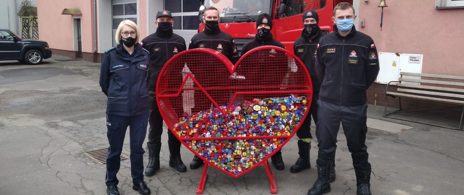 Obraz przedstawia policjantkę i strażaków stojących przy metalowym sercu wewnątrz którego znajdują się plastikowe nakrętki. W tle samochód pożarniczy i budynek komendy.