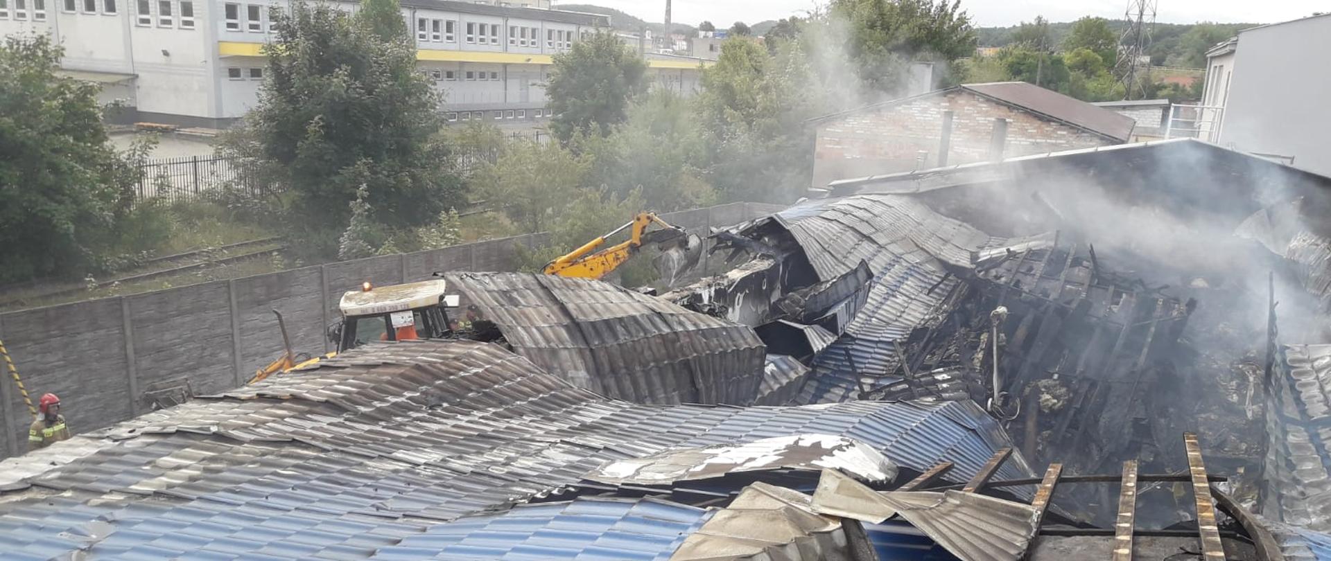 Na zdjęciu widać zapadnięty dach hali magazynowej do wewnątrz budynku. Pracujący ciężki sprzęt podczas usuwania tlącego się materiału palnego na zewnątrz budynku.