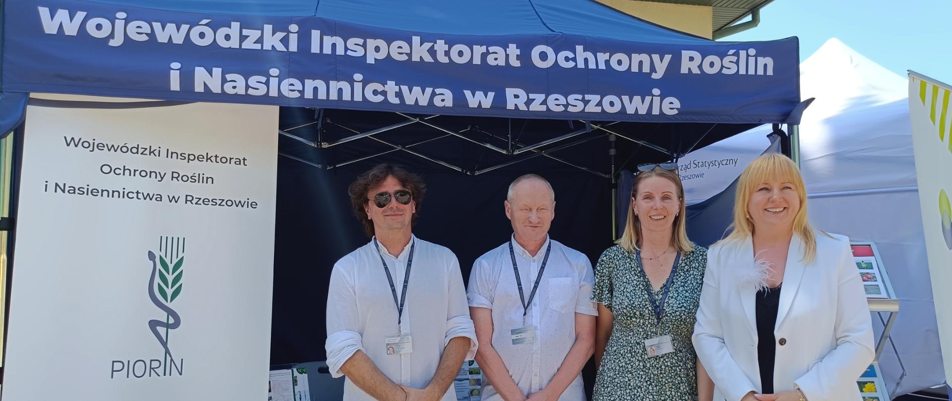 Cztery osoby stoją przed namiotem. Dwie kobiety i dwóch mężczyzn. Po lewej stronie stoi baner z logiem instytucji.