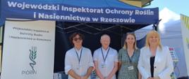 Cztery osoby stoją przed namiotem. Dwie kobiety i dwóch mężczyzn. Po lewej stronie stoi baner z logiem instytucji.
