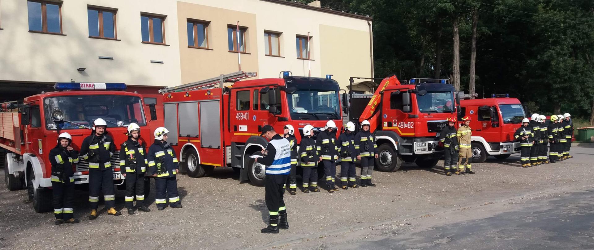 Ćwiczenia powiatowego plutonu ratowniczego. Na placu manewrowym przed jednostką OSP zduny stoją cztery pojazdy pożarnicze. Trzy z nich to pojazdy gaśnicze, natomiast czwarty to pojazd wężowy. Pojazdy zostały ustawione w jednym szyku przodem do wyjazdu. Przed samochodami w szeregu stoją strażacy stanowiący obsady wozów bojowych. Dowódca plutonu sprawdza zgodnie z listą potencjał ratowniczy zgromadzony w miejscu koncentracji. Wszyscy strażacy mają ubrania bojowe i hełmy. W tle widać budynek strażnicy OSP Zduny
