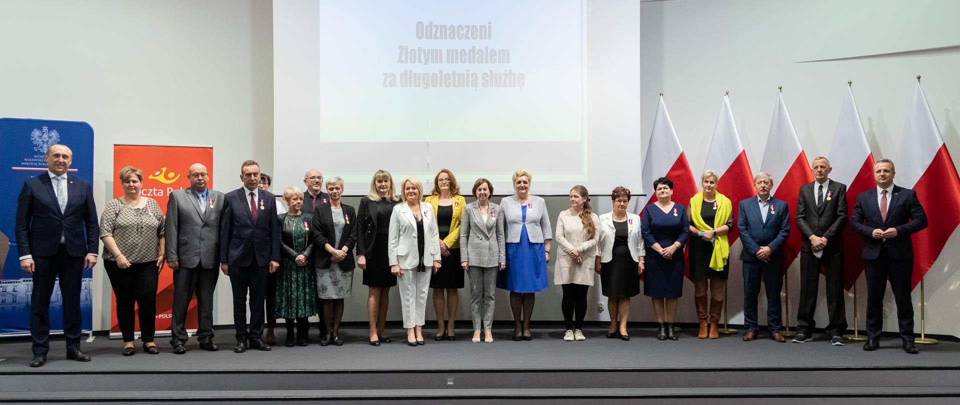 Grupa ludzi stojąca na tle jasnego ekranu, biało-czerwonych flag i bannerów