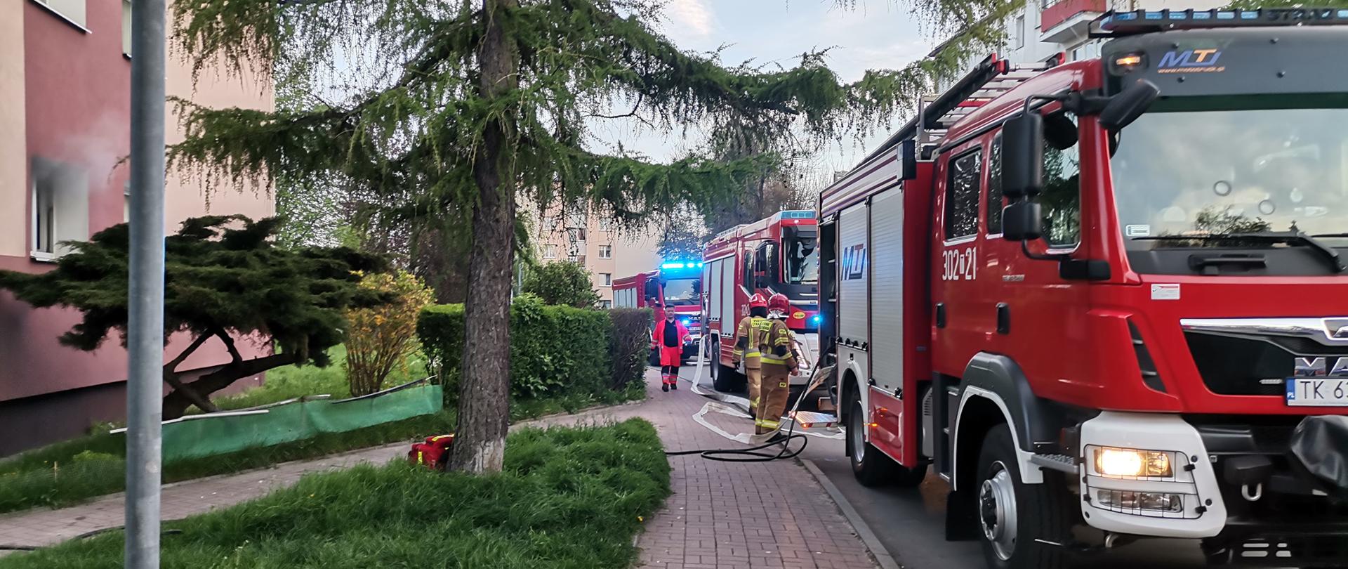 Zdjęcie przedstawia ulicę Urzędniczą przy której zaparkowały trzy samochody pożarnicze. Z lewej strony widać fragment budynku, w którym wybuchł pożar. 
