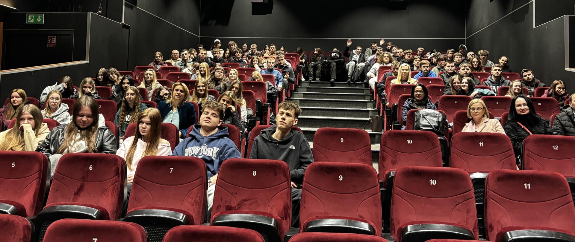 Osoby siedzą na czerwonych fotelach w sali kinowej, wypełniając niemal wszystkie rzędy.