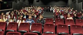 Osoby siedzą na czerwonych fotelach w sali kinowej, wypełniając niemal wszystkie rzędy.