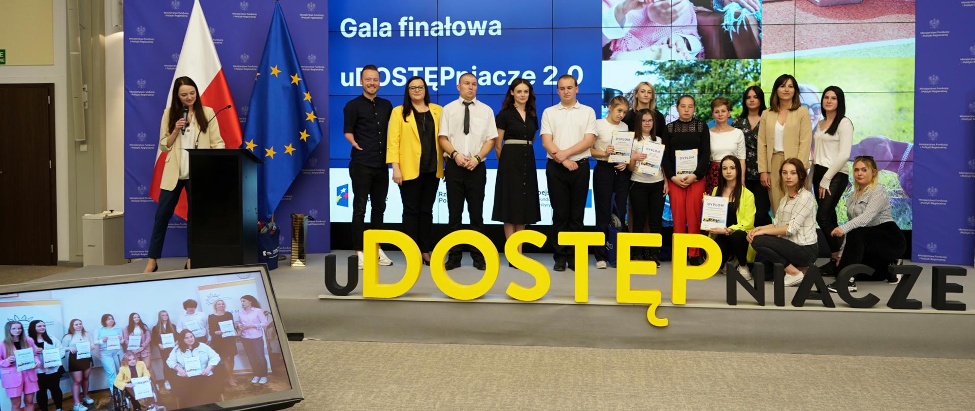W sali konferencyjnej na scenie grupa młodych ludzi. Wśród nich wiceminister Małgorzata Jarosińska-Jedynak. za nimi na ekranie napis Udostępniacze i lustracje osób z niepełnosprawnościami. Z tyłu dwie flagi PL iUE, z przodu monitor.