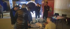 Na zdjęciu widoczni strażacy z Komendy Powiatowej Państwowej Straży Pożarnej w Gorlicach w ubraniach koszarowych czarnych i piaskowych obok sceny 32 Finału Wielkiej Orkiestry Świątecznej Pomocy na hali Ośrodka Sportu i Rekreacji w Gorlicach na stoisku uczą pierwszej pomocy na fantomach osoby zainteresowane tą tematyką.