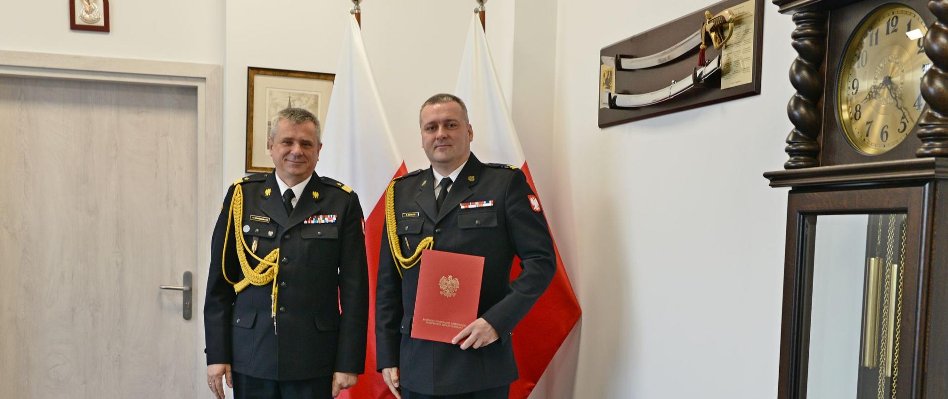 Pomorski komendant wojewódzki Państwowej Straży Pożarnej stoi obok strażaka trzymającego czerwoną teczkę. Za nimi stoją flagi państwowe Polski.