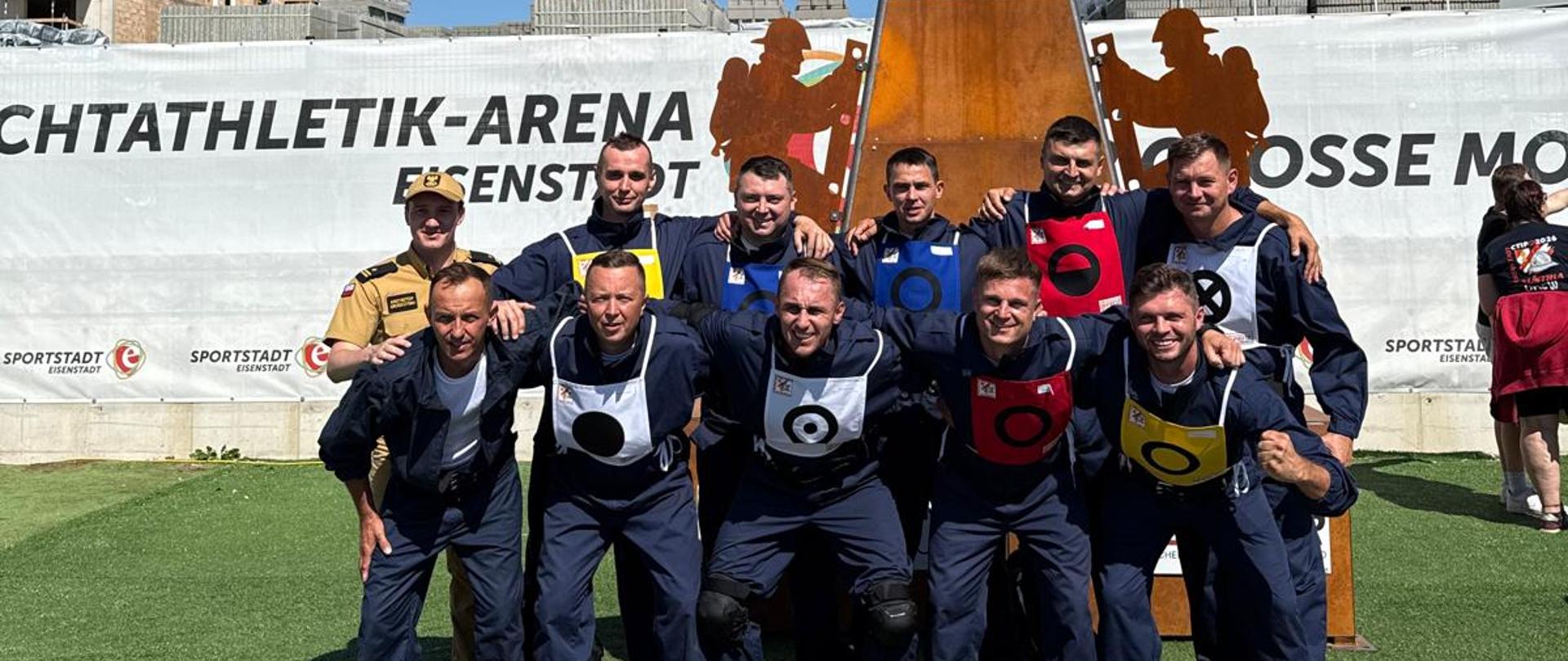 Zdjęcie przedstawia grupę uśmiechniętych strażaków pozujących do wspólnego zdjęcia na trawiastym boisku podczas zawodów CTIF 2026 w Austrii. Dziewięciu mężczyzn w ciemnoniebieskich kombinezonach i kolorowych plastronach startowych stoi w dwóch rzędach, obejmując się ramionami. Przed nimi, na zielonej trawie, ułożony jest równy rząd czerwonych kasków strażackich. W tle widać duży, brązowy znicz olimpijski z logo zawodów, białe banery reklamowe z napisami „LEICHTATHLETIK-ARENA EISENSTADT” oraz plac budowy z rusztowaniami pod bezchmurnym, niebieskim niebem.