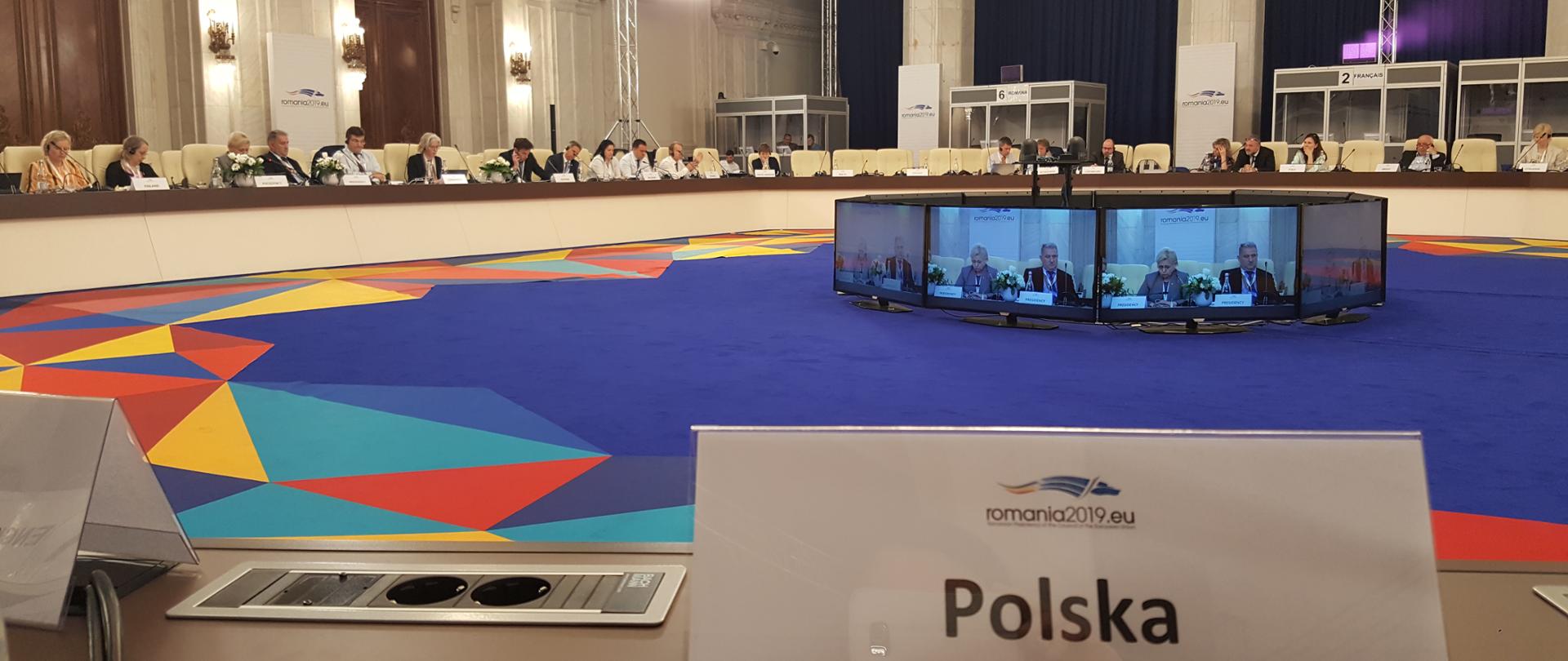 Widok na salę konferencyjną i prelegentów przy okrągłym stole. Na pierwszym planie tabliczka z napisem "Polska".