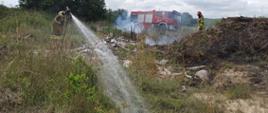 Przedstawia akcję gaśniczą podczas pożaru nielegalnego wysypiska śmieci w pobliżu miejscowości Pomianowo w powiecie białogardzkim. Na zdjęciu rota strażaków w ubraniach specjalnych gasi prądem wodnym stertę śmieci, w głębi zdjęcia samochód strażacki