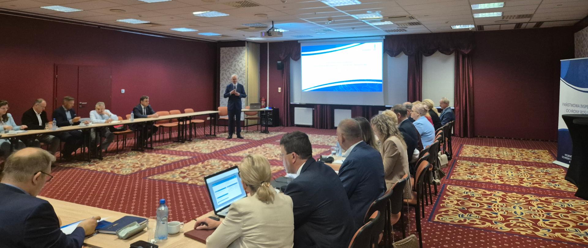 Przestronna sala konferencyjna. Ściany są bordowe, na podłodze bordowy dywan w żółte wzory. Przy jasnych stołach ułożonych w kształcie litery U siedzą zebrani ludzie na wprost mają ekran z wyświetlaczem. Jeden mężczyzna stoi i przemawia. 