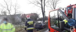Widać czerwony samochód strażacki, w pobliżu jest strażak, w oddali jest uszkodzony pojazd w rowie