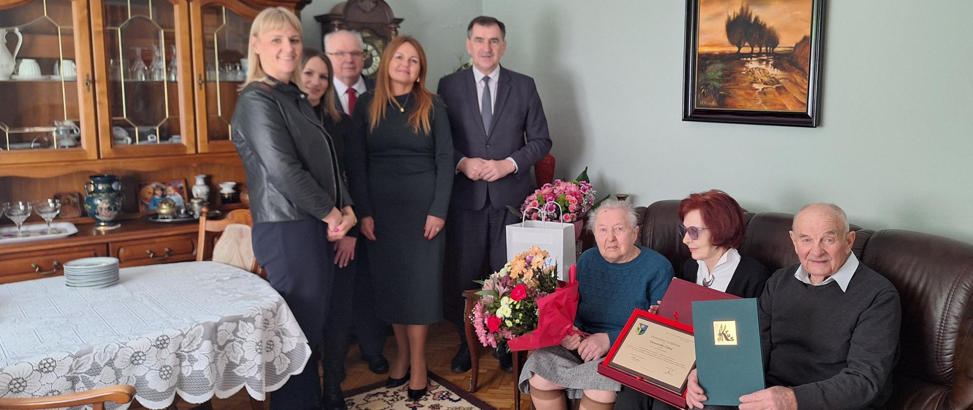 Trzy kobiety i dwóch mężczyzn stoą w pokoju obok obok kanapy. Na kanapie siedzą: starsza kobieta z bukietem kwiatów, obok trochę młodsza kobieta z dyplomem w dłoniach i starszy mężczyzna z zieloną teczką z logo KRUS.