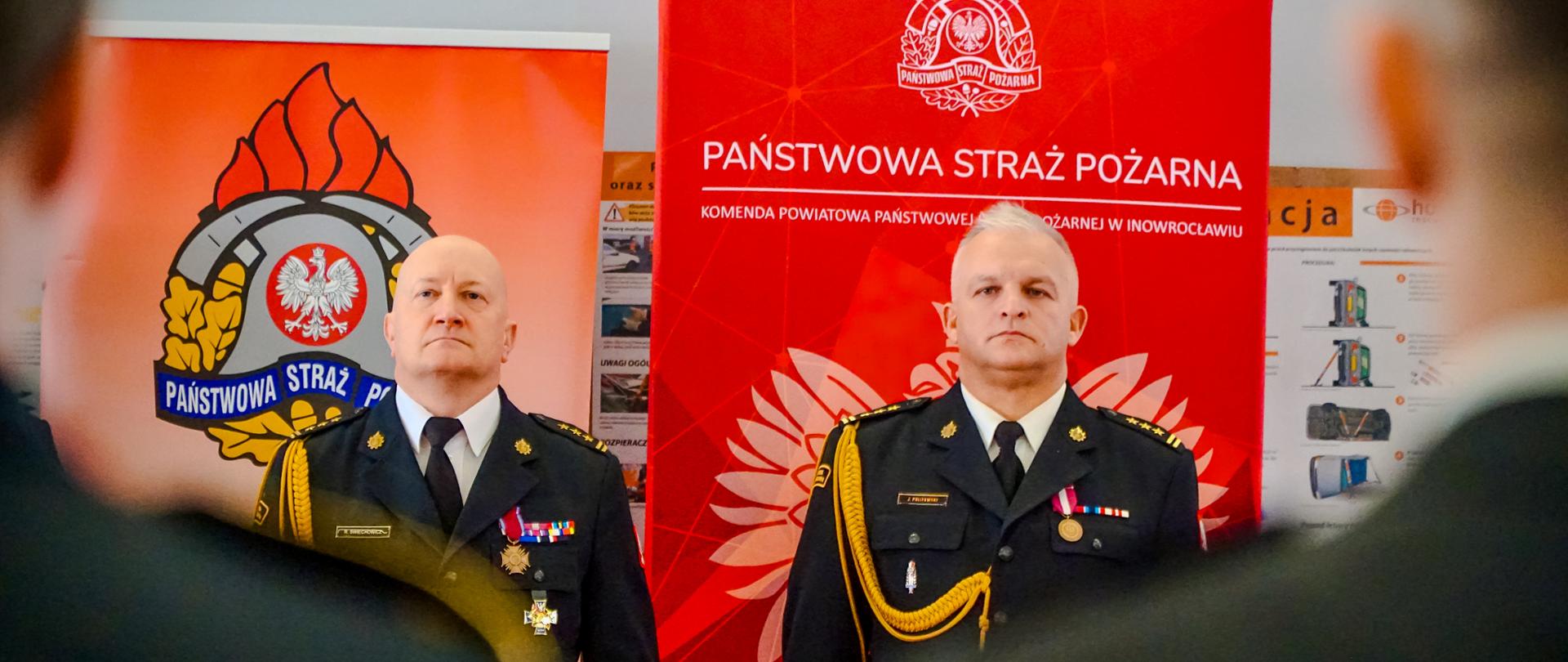 Widok na dwóch oficerów PSP w mundurach galowych. Za nimi banery firmowe. Oni w pozycji zasadniczej. 
