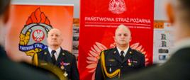 Widok na dwóch oficerów PSP w mundurach galowych. Za nimi banery firmowe. Oni w pozycji zasadniczej. 