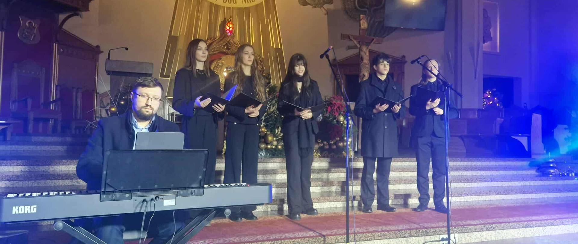 Zespół wokalny oraz pianista występują podczas koncertu kolęd na stopniach prezbiterium kościoła.
