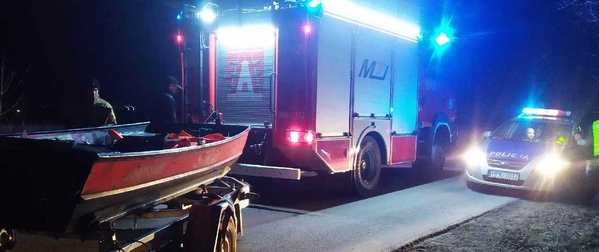 Na zdjęciu po lewej stronie na pierwszym planie widać łódź na przyczepie. Przyczepa zamocowana jest do strażackiego samochodu gaśniczego. Po prawej stronie widać radiowóz policyjny. Zdjęcie zrobione w nocy.