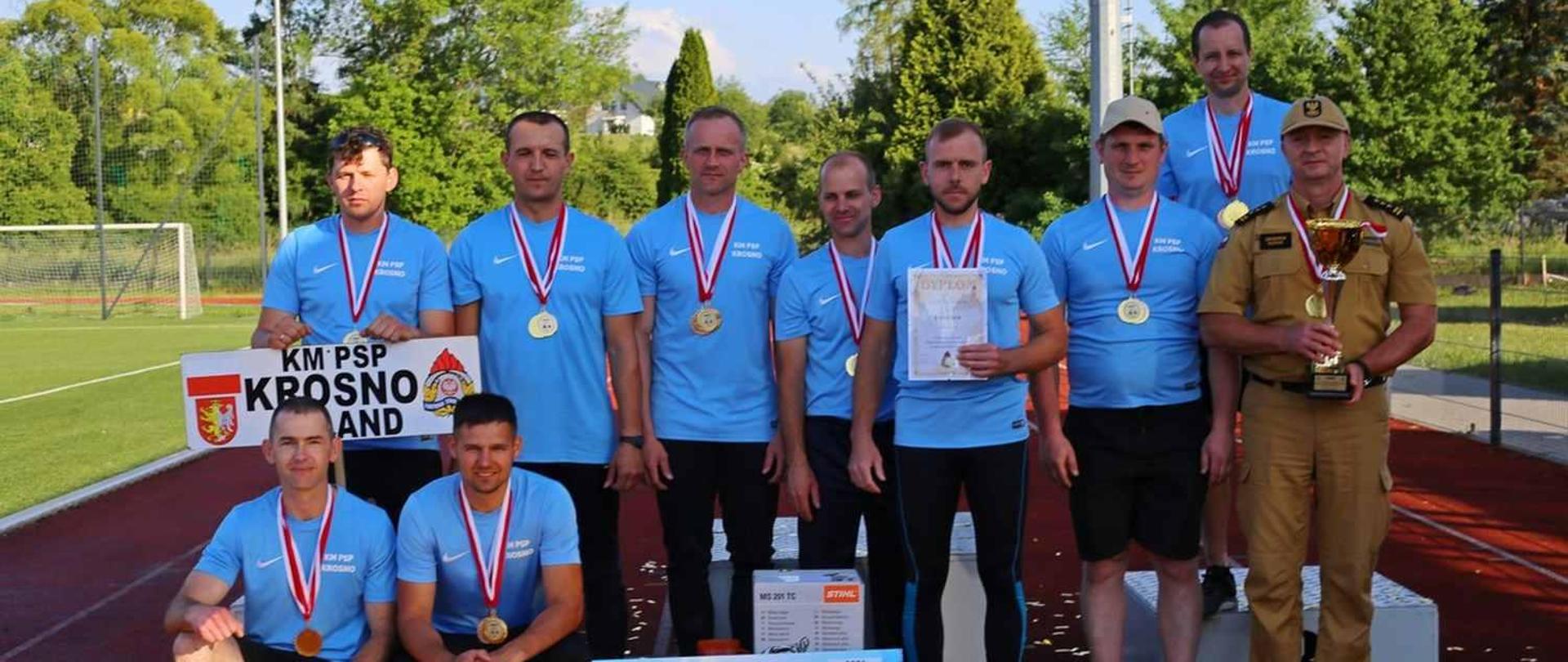 Zdjęcie przedstawia reprezentację KM PSP Krosno podczas zawodów w sporcie pożarniczym. Wraz z drużyną stoi Komendant Miejski PSP w Krośnie, który trzyma puchar za zajęcie I miejsca w klasyfikacji generalnej zawodów. Na szyi każdego z zawodników wisi złoty medal.