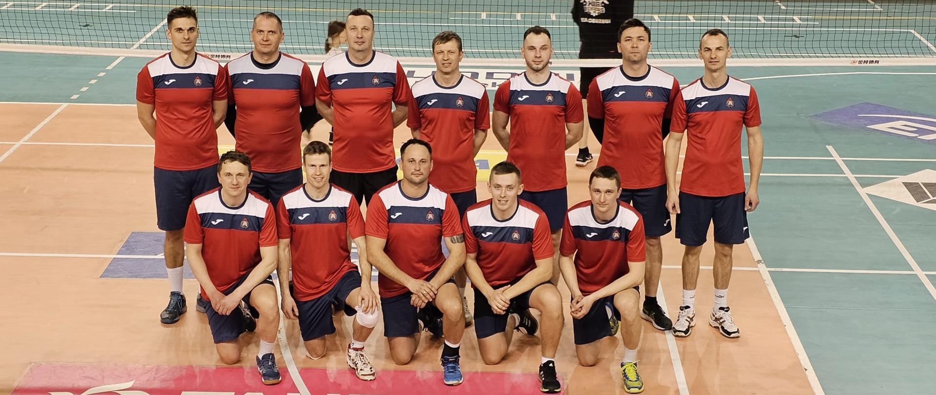 Poziome, pełnokolorowe zdjęcie przedstawia drużynę siatkarzy Komendy Powiatowej Państwowej Straży Pożarnej w Przysusze pozujących do zdjęcia na boisku sportowym.
W pierwszym rzędzie, na dole zdjęcia, klęczy pięciu mężczyzn. W drugim rzędzie, za nimi, stoi siedmiu mężczyzn. Wszyscy ubrani są w identyczne stroje sportowe: czerwono-granatowe koszulki z białymi wstawkami i granatowe spodenki. Na lewej piersi koszulek widnieje logo.
Mężczyźni mają krótkie fryzury i patrzą prosto w obiektyw. Tło zdjęcia stanowi boisko do siatkówki z białymi liniami i fragmentem siatki widocznym na górze kadru. Na podłodze boiska widoczne są również reklamy sponsorów, w tym wyraźne logo "TAURON" na pierwszym planie. Oświetlenie w sali jest jasne. W tle, za siatką, widać fragment postaci innej osoby. Cała scena sugeruje, że zdjęcie zostało wykonane po meczu siatkówki.
