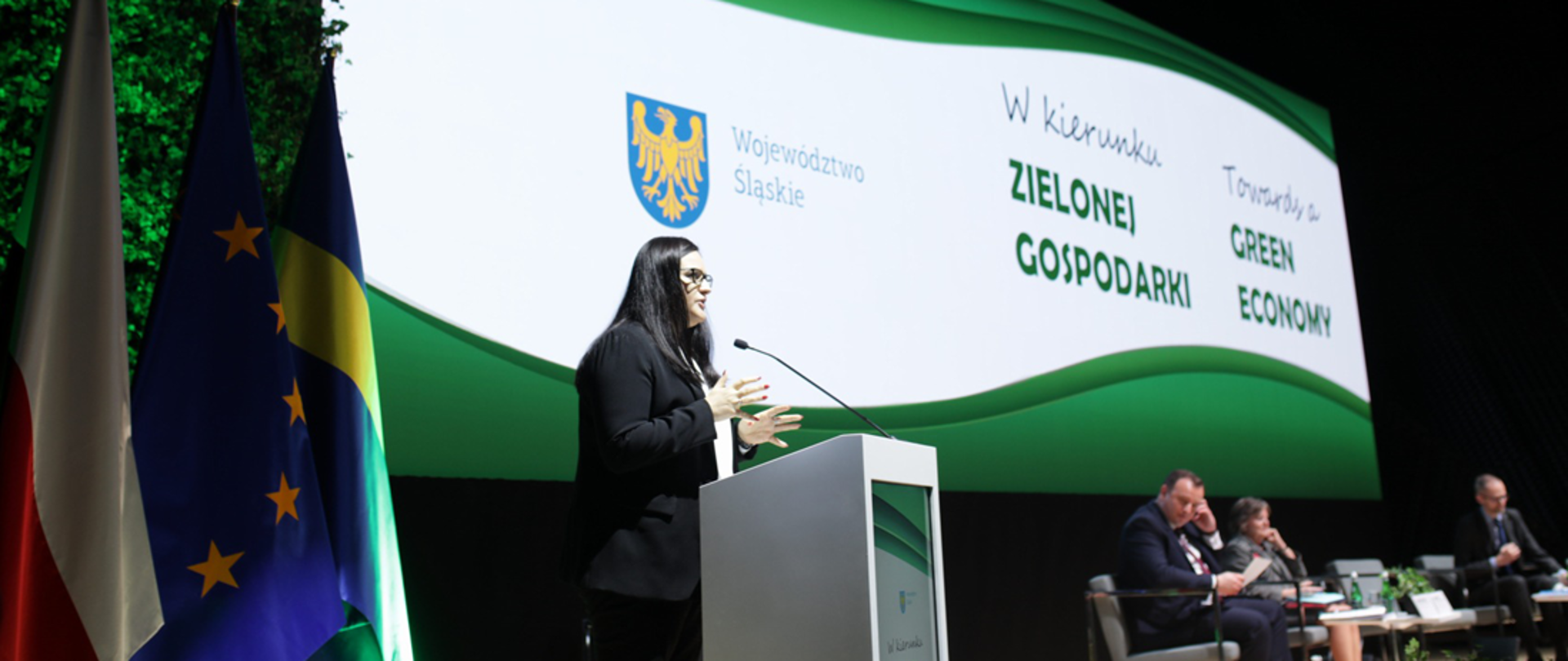 Na zdjęciu od lewej stoją trzy flagi. Dalej mównica z mikrofonem, w której przemawia minister Jarosińska-Jedynak. Z prawej strony siedzą na fotelach trzy osoby. Z tyłu baner biało-zielony z napisami "w kierunku zielonej gospodarki" oraz godło województwa śląskiego.